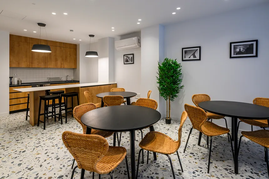Cocina comunitaria de coliving con muebles de madera clara, una isla central con encimera blanca y dos mesas de comedor redondas negras con sillas de mimbre de color claro.