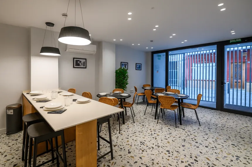 Comedor compartido de un coliving con mesas redondas y sillas de mimbre, mesa alta con vajilla preparada, iluminación cálida y ventanales que dan a la calle.