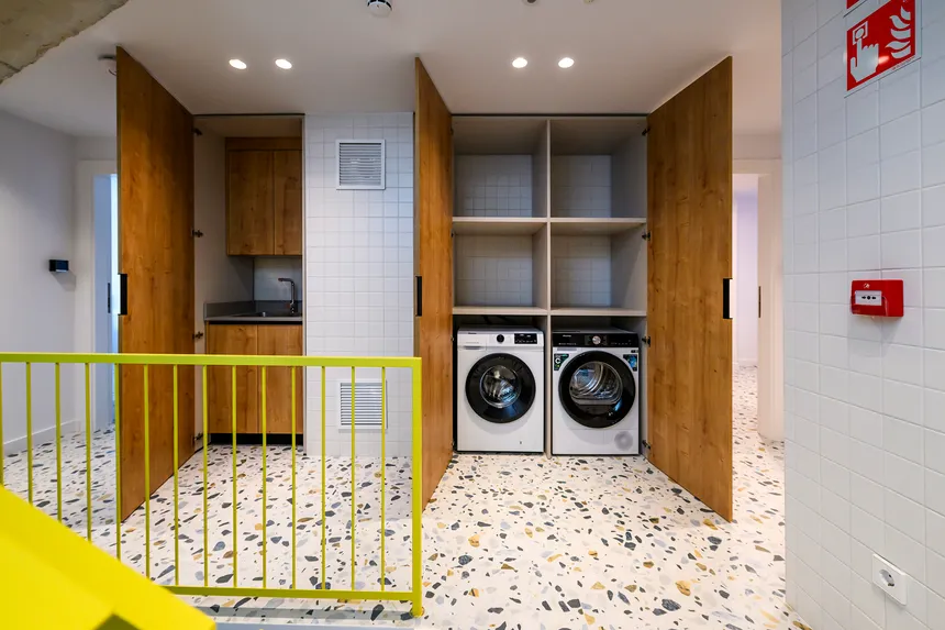 Lavandería de coliving con lavadora y secadora integradas en un mueble de madera, estanterías abiertas y un pequeño fregadero a la izquierda.