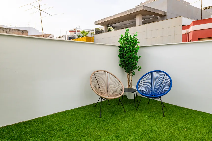 Terraza con césped artificial, dos sillas circulares de diseño en beige y azul, una mesa auxiliar negra y una planta decorativa.