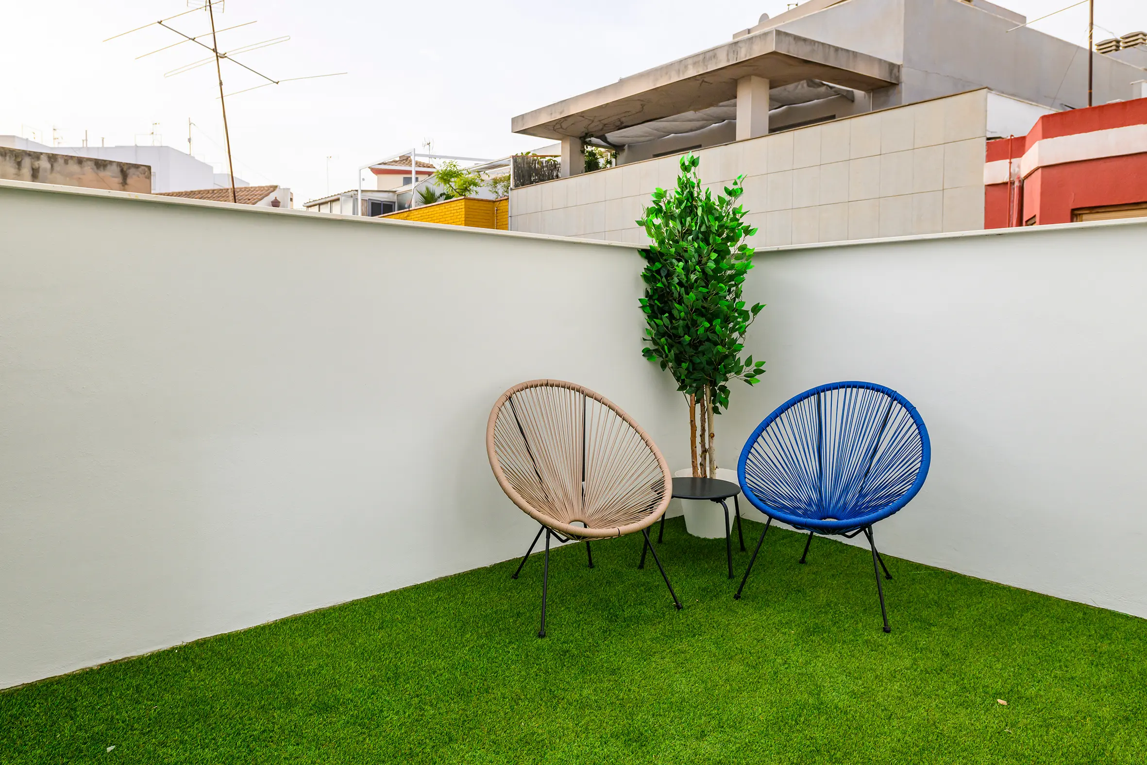 Terraza con césped artificial, dos sillas circulares de diseño en beige y azul, una mesa auxiliar negra y una planta decorativa.